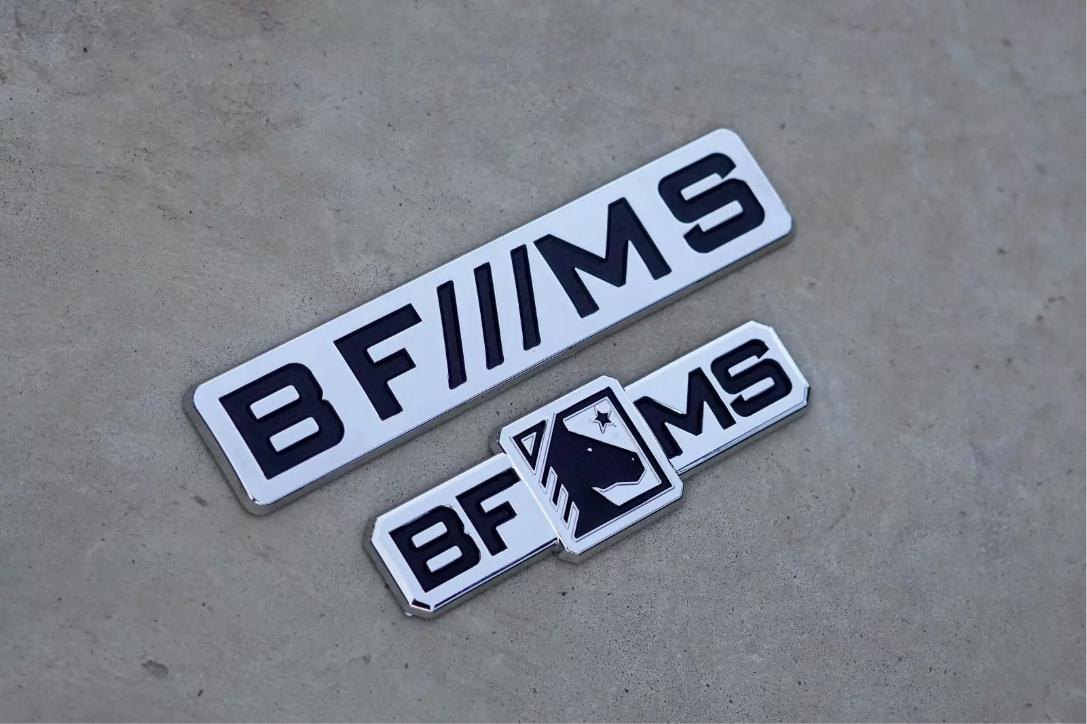 Dois emblemas cromados BF///MS com fundo branco e letras pretas em relevo, um deles com o brasão do cavalo, sobre piso de concreto