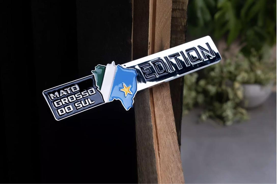 Emblema Mato Grosso do Sul Edition, com o mapa do estado em azul e a estrela amarela, apoiado em madeira