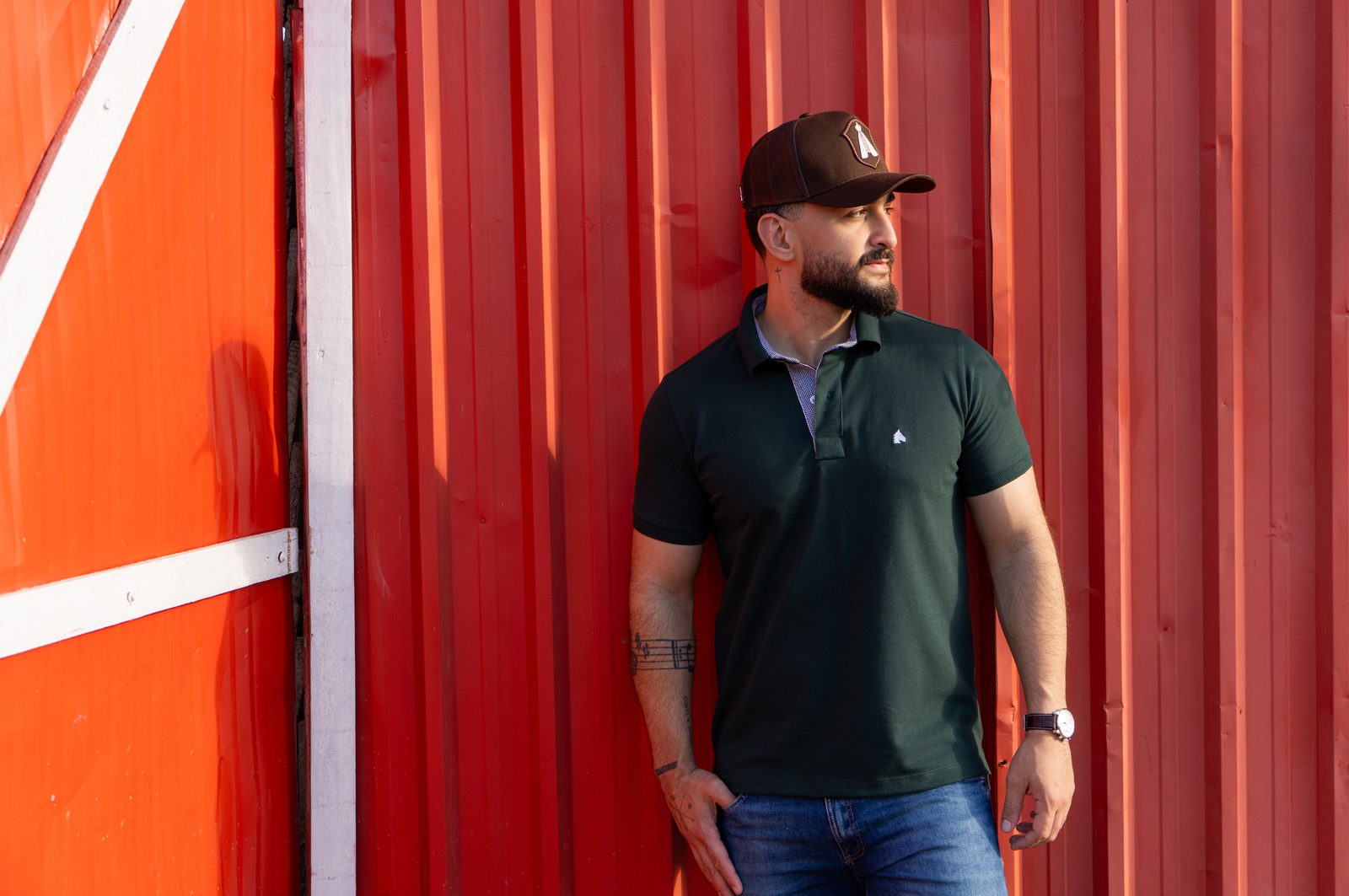 Homem usando polo verde e boné trucker marrom da BF///MS, apoiado em um container vermelho sob luz de fim de tarde