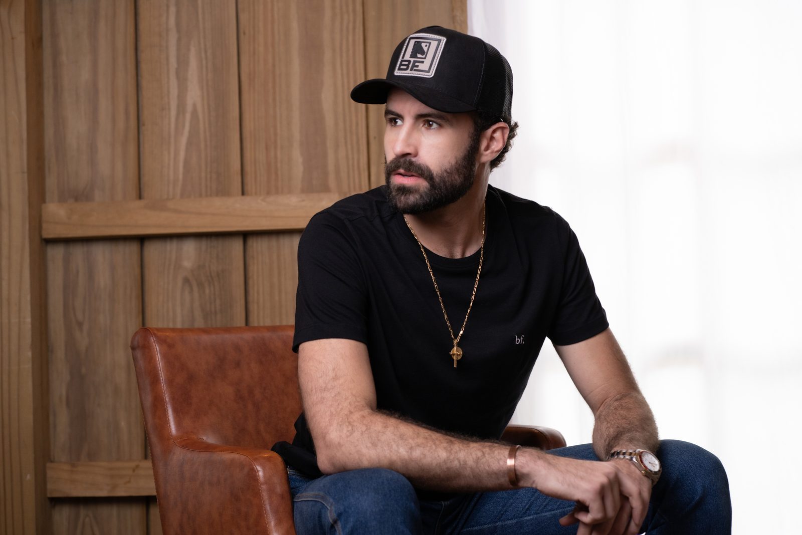Homem sentado em poltrona de couro usando camiseta preta com logo bf. e boné trucker preto da BF///MS, com parede de madeira ao fundo